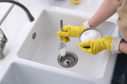 A person wearing gloves pours a spoonful of baking soda into a drain.