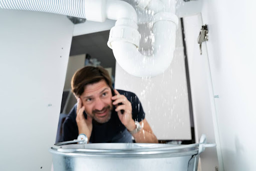 A man on the phone as he looks at a plumbing leak under a sink.